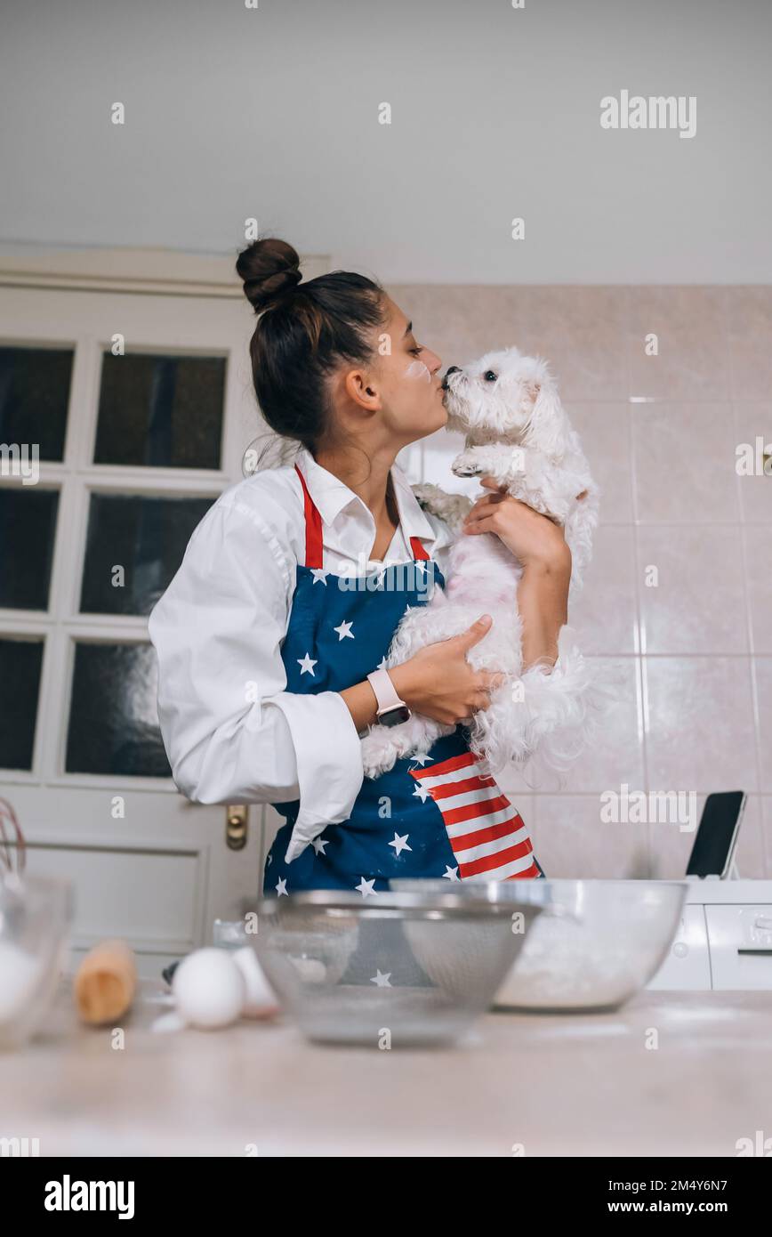 Junge Frau in der Küche küsst einen niedlichen weißen maltesischen Hund Stockfoto