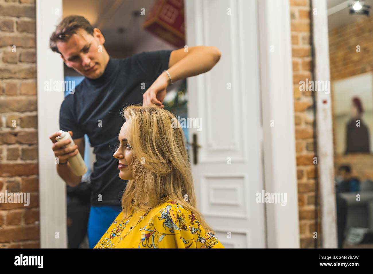 Experte männliche Stylistin, die blondes Haar seiner jungen, erwachsenen Klientin stylt. Friseurinnenraum. Unscharfer Hintergrund, Kopierbereich. Hochwertiges Foto Stockfoto