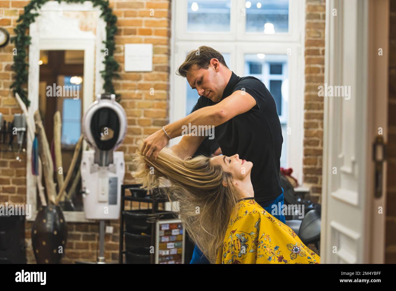 Profi-Friseurin während der Arbeit. Männlicher Friseur schneidet blonde Haare seines Klienten auf unkonventionelle Weise. Friseurinnenraum. Speicherplatz kopieren. Hochwertiges Foto Stockfoto