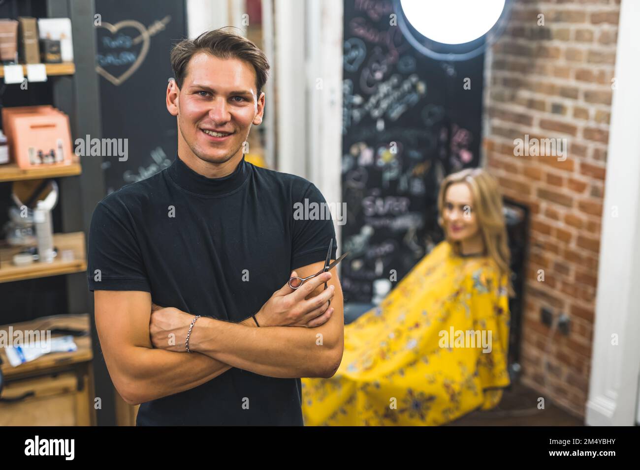 Junger erwachsener, männlicher Haarkünstler, der mit einem Lächeln in die Kamera schaut. Blonde weibliche Klientin im Hintergrund. Friseurinnenraum. Hochwertiges Foto Stockfoto
