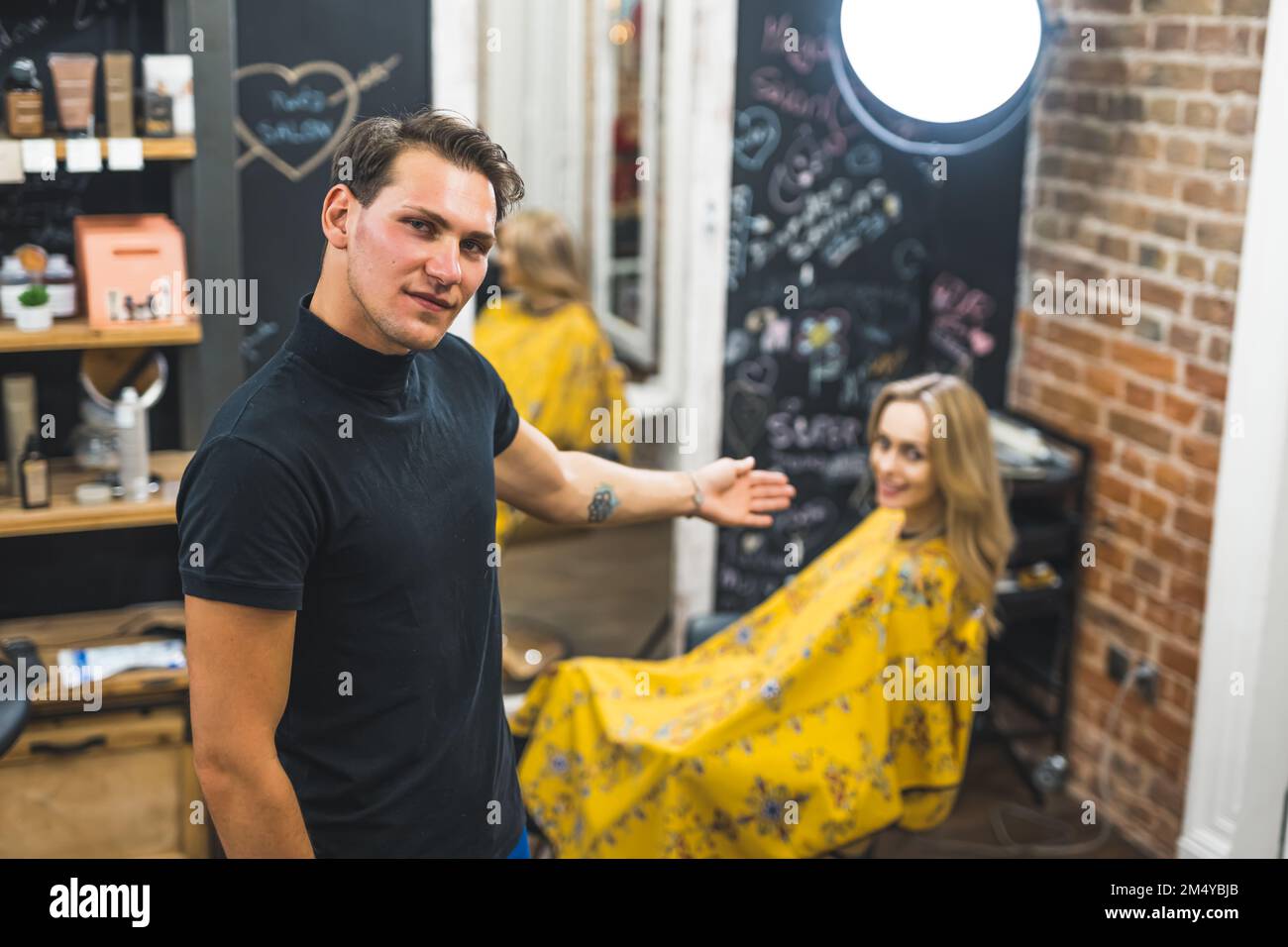 Stolzer, männlicher Friseur, der die neue Frisur seines Klienten vor der Kamera zeigt. Unscharfe Mandantin auf Friseurstuhl im Hintergrund. Hochwertiges Foto Stockfoto