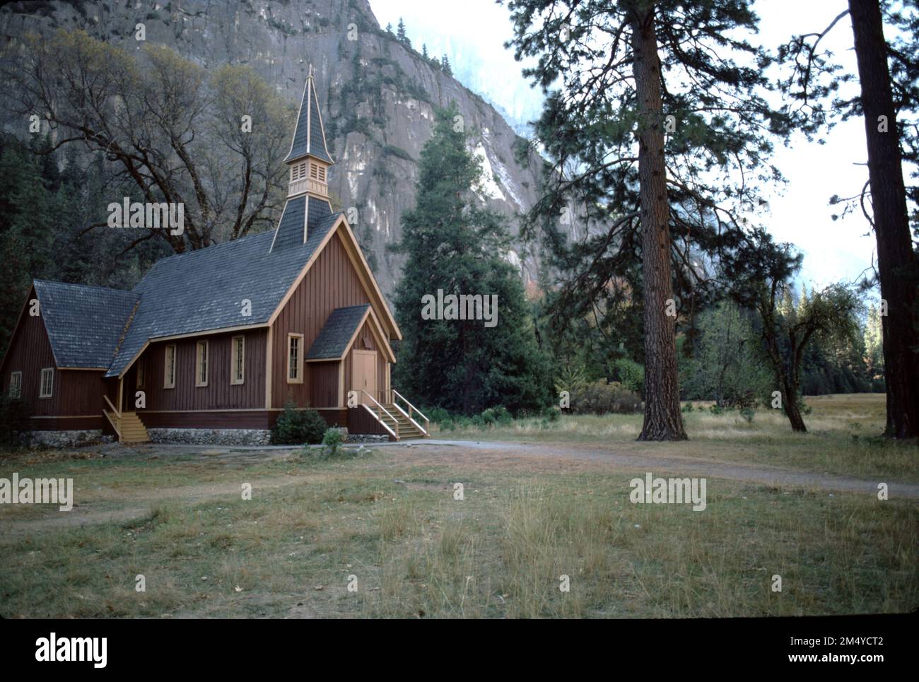 Yosemite-Nationalpark CA USA. 9/1984. Merced River Bride und die Yosemite Valley Chapel. Die Ahwahnee Bridge wurde 1928 über den Merced mit drei Bögen gebaut, von denen einer 42 Fuß (13 m) und die anderen 39 Fuß (12 m) groß sind und eine Gesamtlänge von 122 Fuß (37 m) haben. Die Brücke ist 39 Fuß (12 m) breit mit einer 27 Fuß (8,2 m) langen Straße, einem 5 Fuß (1,5 m) langen Gehweg und einem 7 Fuß (2,1 m) langen Weg. Sie führt die Mirror Lake Road, die einen Blick auf Half Dome für den Verkehr in Richtung Osten bietet. Es kostete 59.913,09 Dollar. Die Kapelle wurde 1879 unter der Schirmherrschaft der Yosemite Union Chapel Association erbaut. Stockfoto