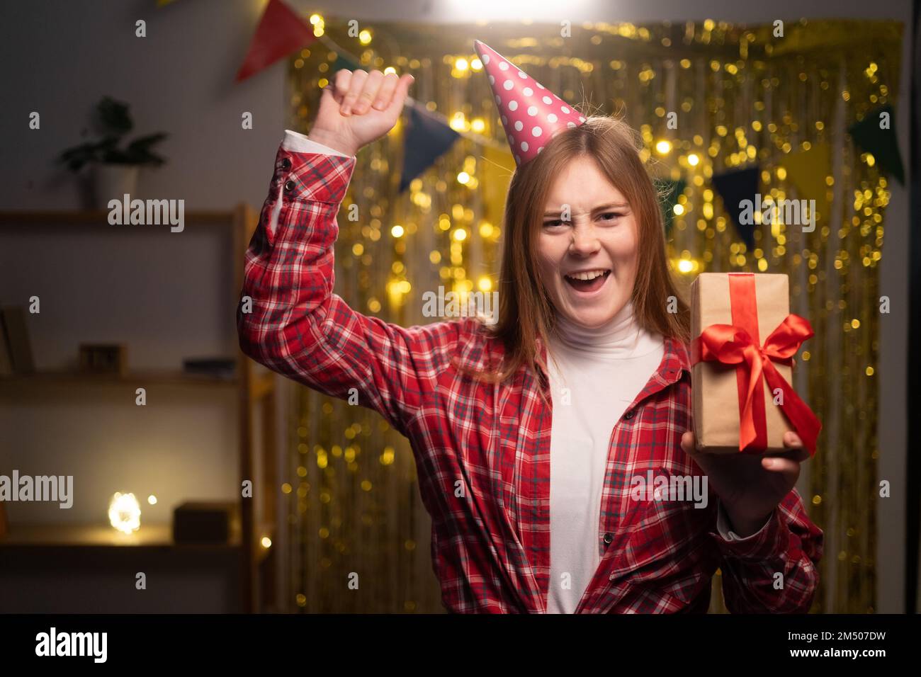 Eine ekstatische, emotionale junge Frau mit Hut und Hemd, die vor Aufregung schreit auf der Party, eine Geschenkbox in der Hand hält, die Faust zusammendrückt, die Geburt feiert Stockfoto