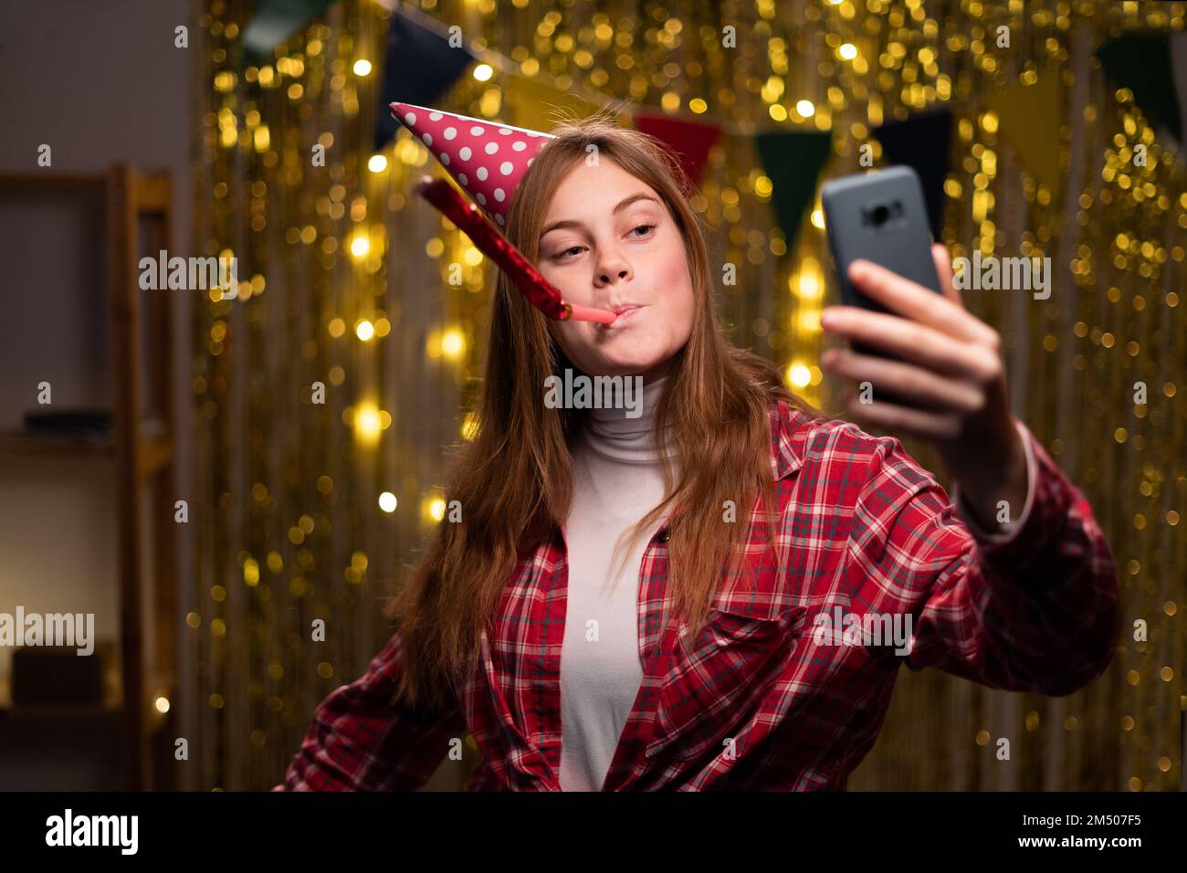 Glückliches Mädchen mit Partylöcher, das in das Partyhorn bläst und ein Mobiltelefon hält, Selfie macht oder einen Videoanruf anlässlich ihres Geburtstags zu Hause macht. Speicherplatz kopieren Stockfoto