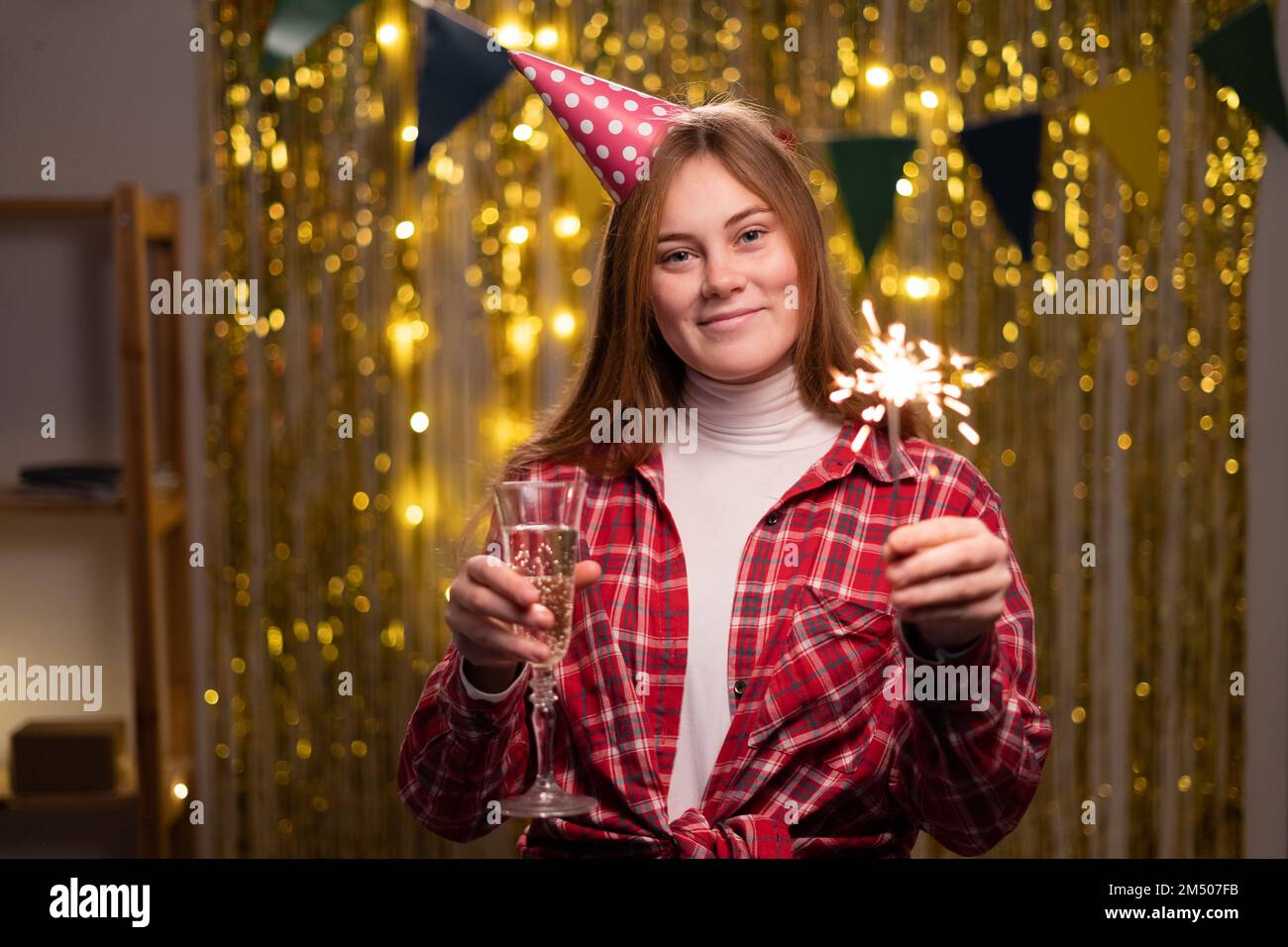 Fröhliche junge Frau mit Konus-Partyhut mit leuchtenden Glitzern und Champagner in den Händen während der Geburtstagsfeier zu Hause. Speicherplatz kopieren Stockfoto