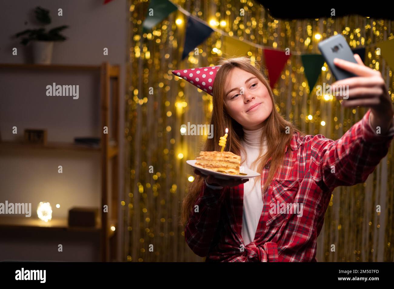 Lächelnde junge Frau in lässigem Pullover Geburtstagsmütze, die abends zu Hause mit Dekor feiert. Geburtstagsparty, Menschen-Emotionen-Konzept. Feiertag Stockfoto