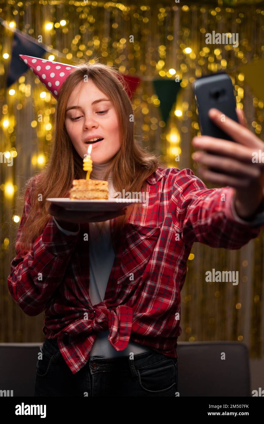 Lächelnde junge Frau in lässigem Hemd Geburtstagsmütze zu Hause feiern. Geburtstagsparty, Menschen-Emotionen-Konzept. Brennende Kerze ausblasen bei C. Stockfoto
