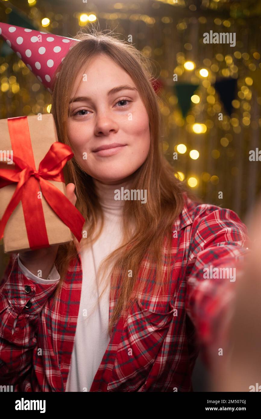 Wunderschöne junge Frau im Hemd, die Selfie mit Geburtstagsgeschenk macht. Hübsche, langhaarige Dame mit einem Geburtstagsgeschenk auf dekoriertem Hintergrund und Taki Stockfoto