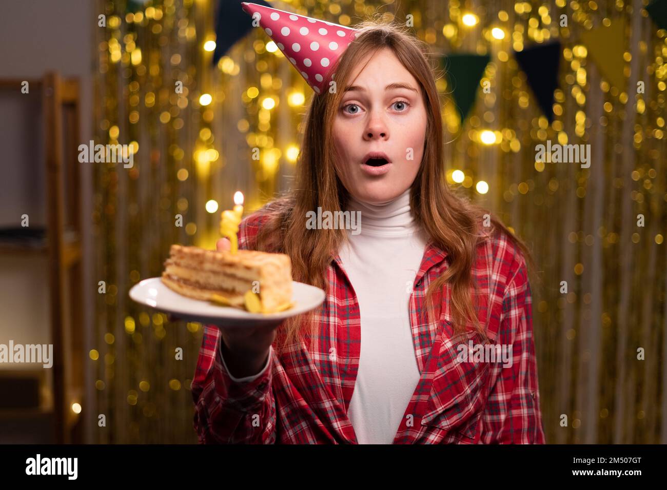 Eine überraschte junge Frau mit einem Hut auf dem Kopf feiert Geburtstag unter Freunden, verblüfft von einem unerwarteten Geschenk. Speicherplatz kopieren Stockfoto
