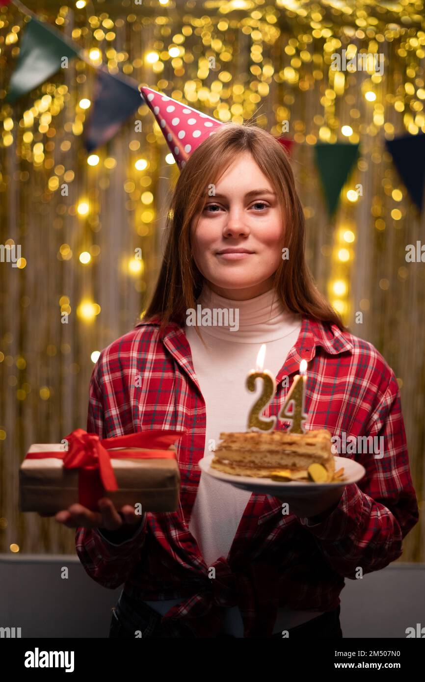 Geburtstagsfeier. Eine junge Frau mit einem Teller mit leckerem Geburtstagskuchen und einem Geschenk im Partyhintergrund. Zum 24. Geburtstag. Speicherplatz kopieren Stockfoto