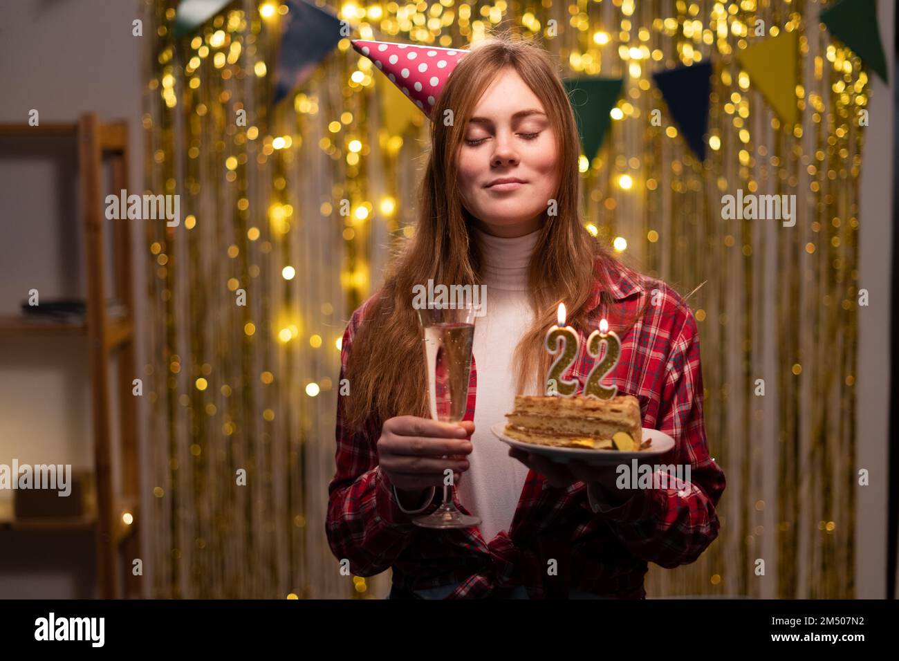 Geburtstagsfeier. Ein weißes Mädchen wünscht sich etwas, indem sie die Augen schließt und einen Geburtstagskuchen mit Kerzen Nummer 22 in der Hand hält. Speicherplatz kopieren Stockfoto
