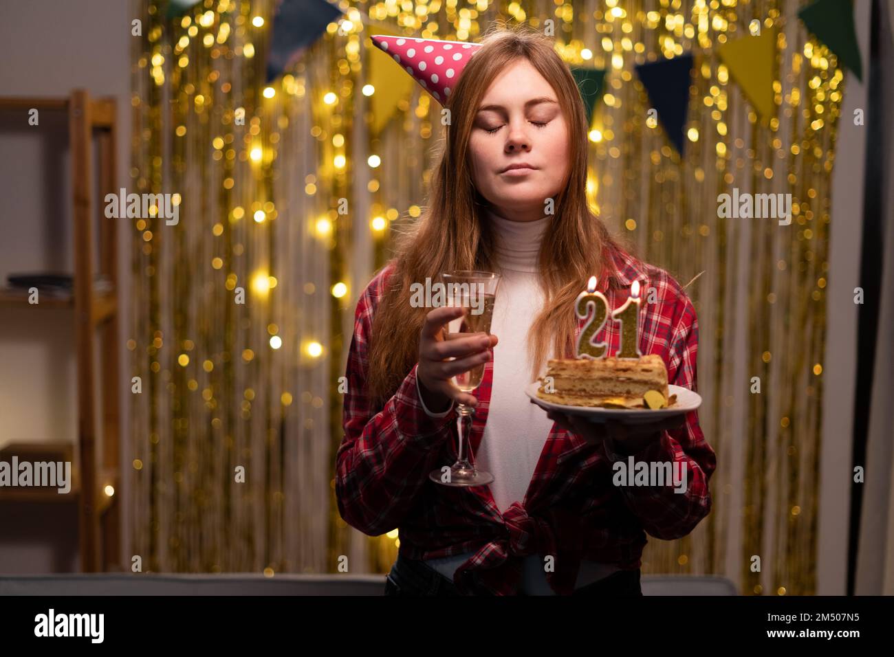 Ein weißes Mädchen wünscht sich etwas, indem sie die Augen schließt und einen Geburtstagskuchen mit Kerzen Nummer 21 in der Hand hält. Speicherplatz kopieren Stockfoto