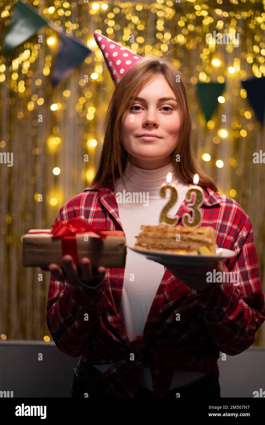 Geburtstagsfeier. Ein Mädchen mit Partyhut, das Kuchen mit Kerzen Nummer 23 hält und ein Geburtstagsgeschenk, das in die Kamera schaut. Speicherplatz kopieren Stockfoto