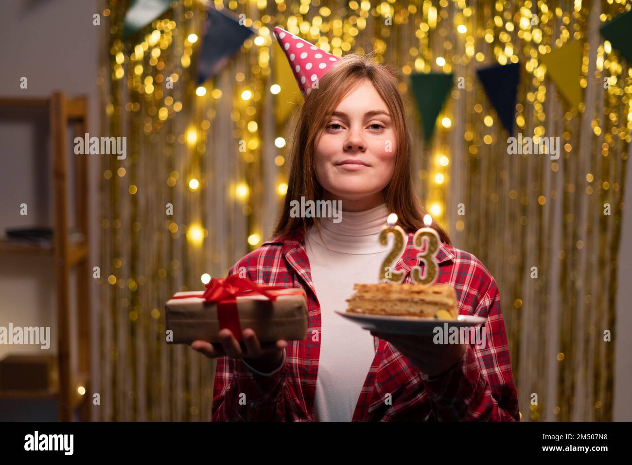 Eine junge Frau mit einem Teller mit leckerem Geburtstagskuchen und einem Geschenk im Partyhintergrund. Zum 23. Geburtstag. Speicherplatz kopieren Stockfoto