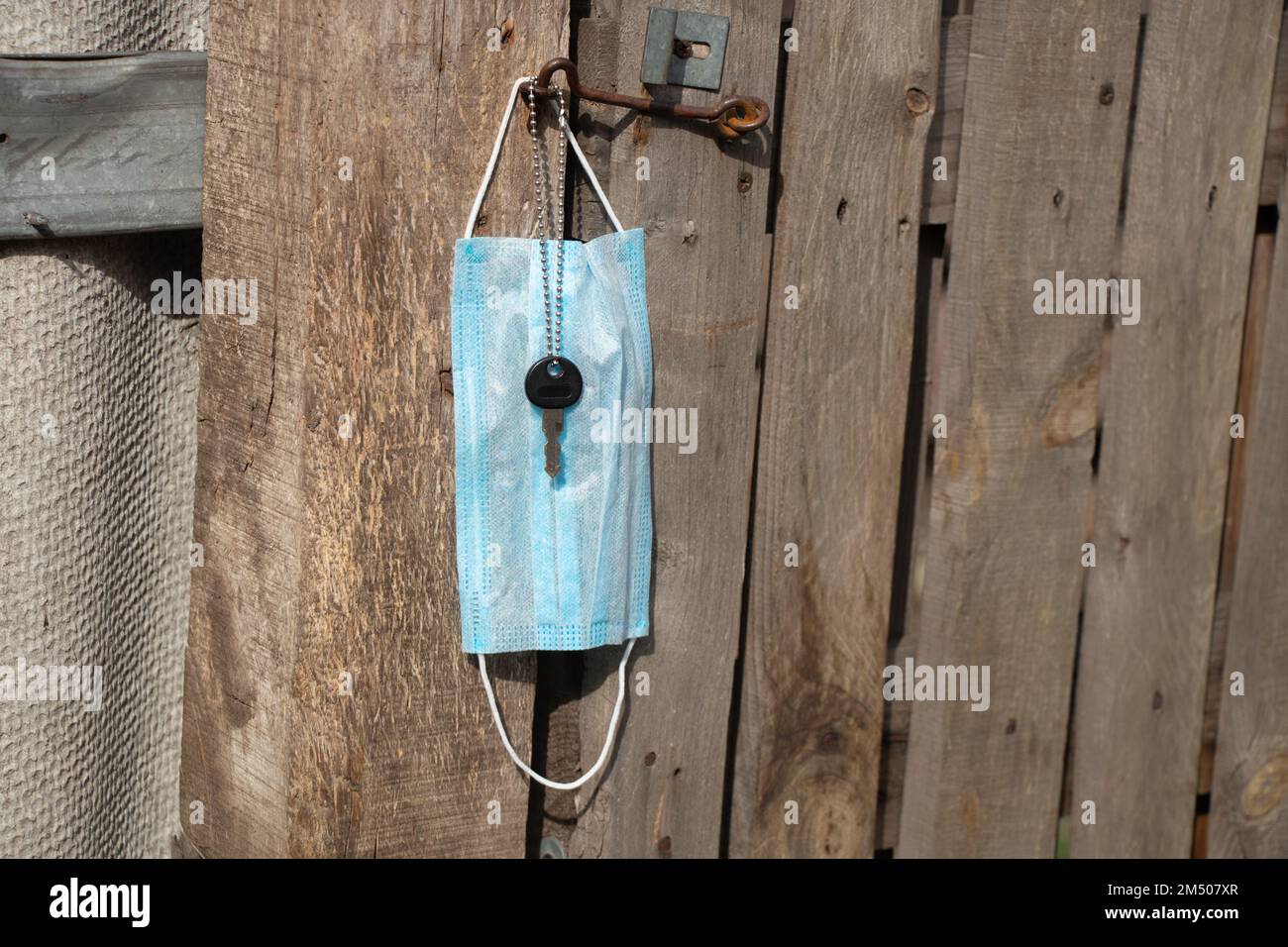 Medizinische Maske hängt an einem Haken und einem Türschlüssel an einem alten Holztor in der Ukraine auf der Straße währenddessen Stockfoto