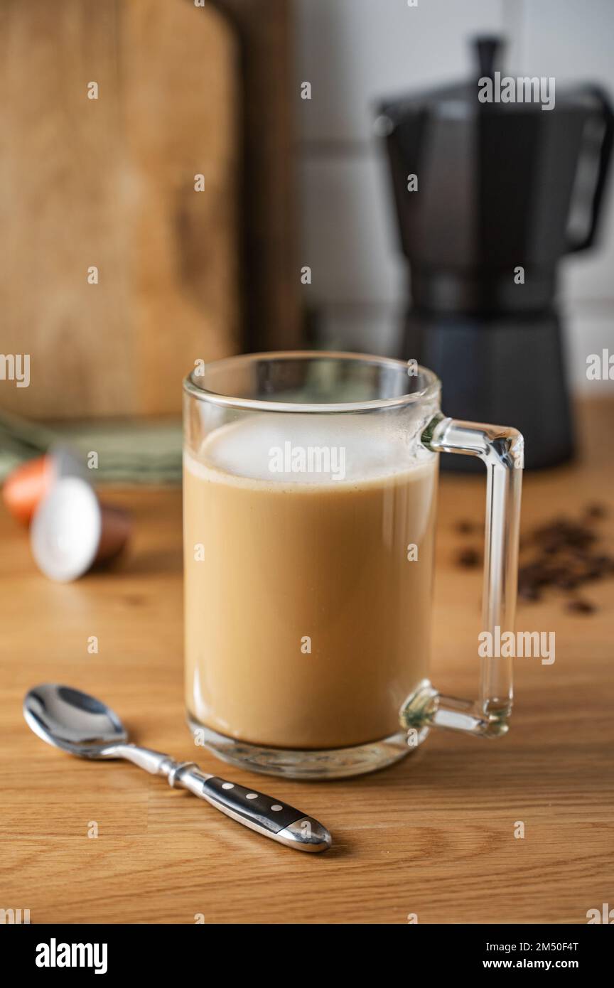 Tasse frisch gebrühten aromatischen Kaffees mit Milch auf dem Küchentisch mit einer Kaffeekanne und verstreuten Körnern aus nächster Nähe. Das Konzept eines frühen Morgens Stockfoto