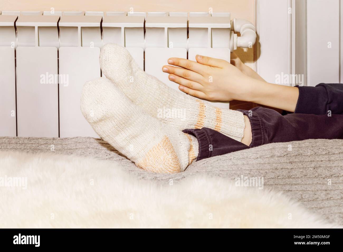 Winter, Heizsaison. Ein Teenager in warmen Kleidern und Stricksocken, der sich in der Nähe des Heizkörpers erwärmt. In der Nähe des Kühlers im Kühlhaus warm halten. Schlechte Hitze Stockfoto