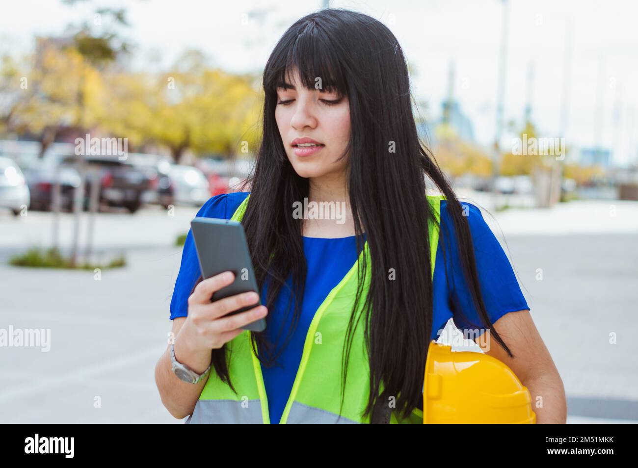 Junge Ingenieurin, die auf der Straße steht und sich ausruht, den Schutzhelm in der Hand hält und ihr Handy benutzt, das Handy beobachtet und tippt, s Stockfoto