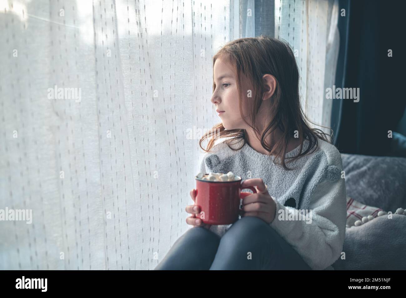 Ein süßes Mädchen auf dem Fensterbrett hält eine rote Tasse Kakao und Marshmallow und schaut aus dem Fenster. Stockfoto
