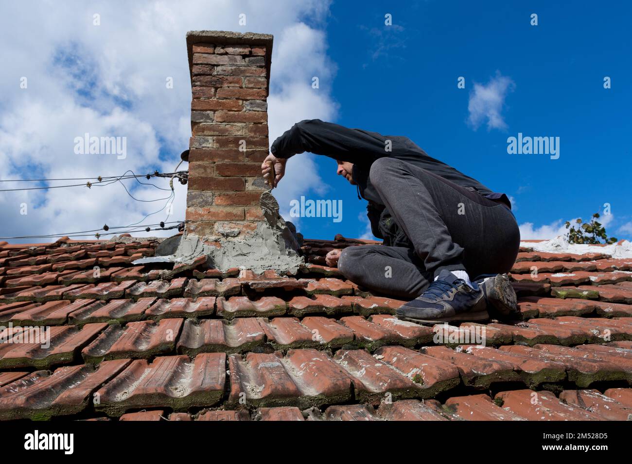 Ein erfahrener Handwerker auf dem Dach eines alten Hauses, der eine Kelle benutzt, um den Kamin zu reparieren. Der Mann sitzt konzentriert auf die anstehende Aufgabe. Himmel im Hintergrund Stockfoto