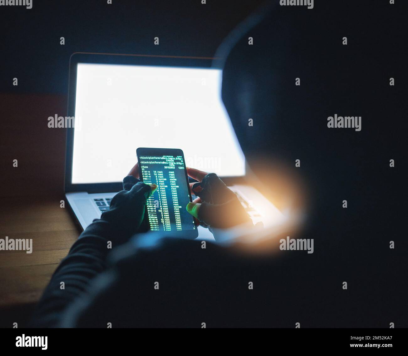 In der schattigen Welt des Online-Hacking. Ein nicht erkennbarer Hacker, der im Dunkeln ein Handy und einen Laptop benutzt. Stockfoto