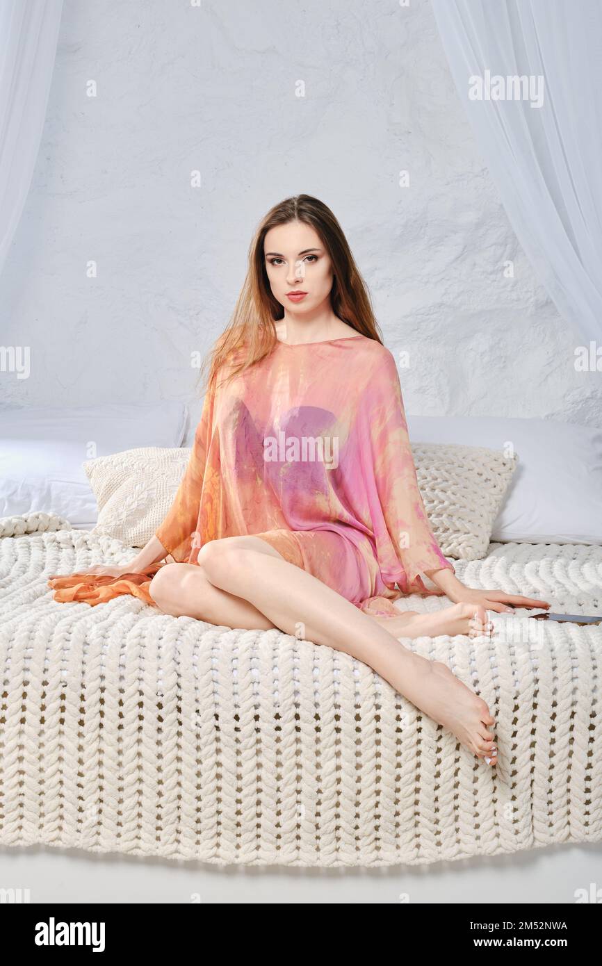 Eine ruhige junge Frau im Seidenmantel, die auf dem Bett sitzt. Das Konzept von Komfort, Behaglichkeit und Entspannung zu Hause. Stockfoto