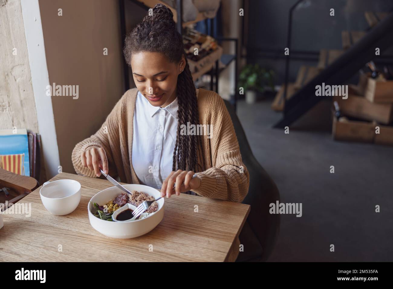 Eine junge afroamerikanische, wunderschöne Frau isst in einem Restaurant zu Mittag und probiert ein gesundes Gericht, serviert in einer weißen Schüssel Stockfoto