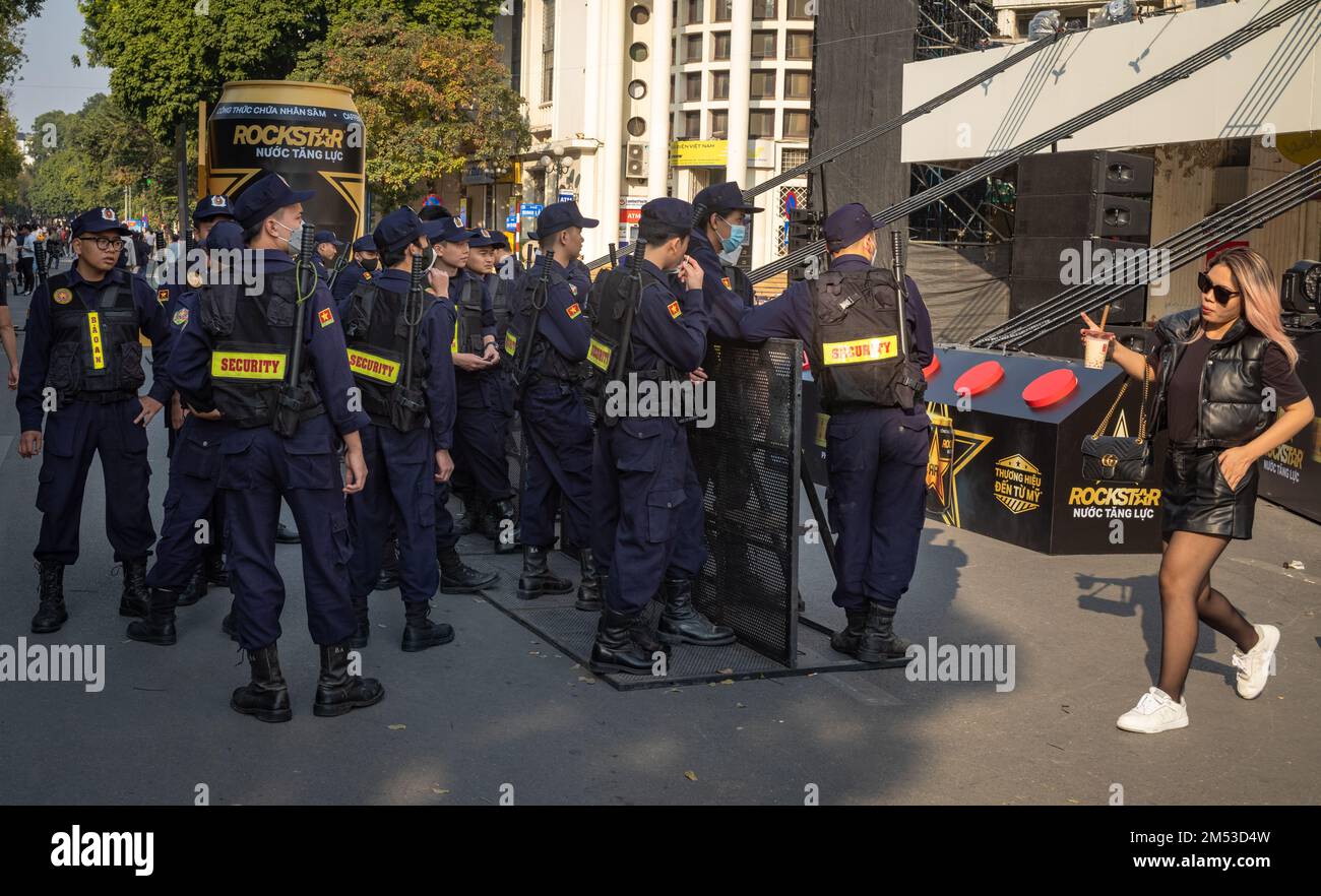 Eine Gruppe privater Sicherheitskräfte mit langen Schlagstöcken wartet auf eine Open-Air-Vorstellung, während eine junge Frau in Hanoi, Vietnam, vorbeiläuft Stockfoto