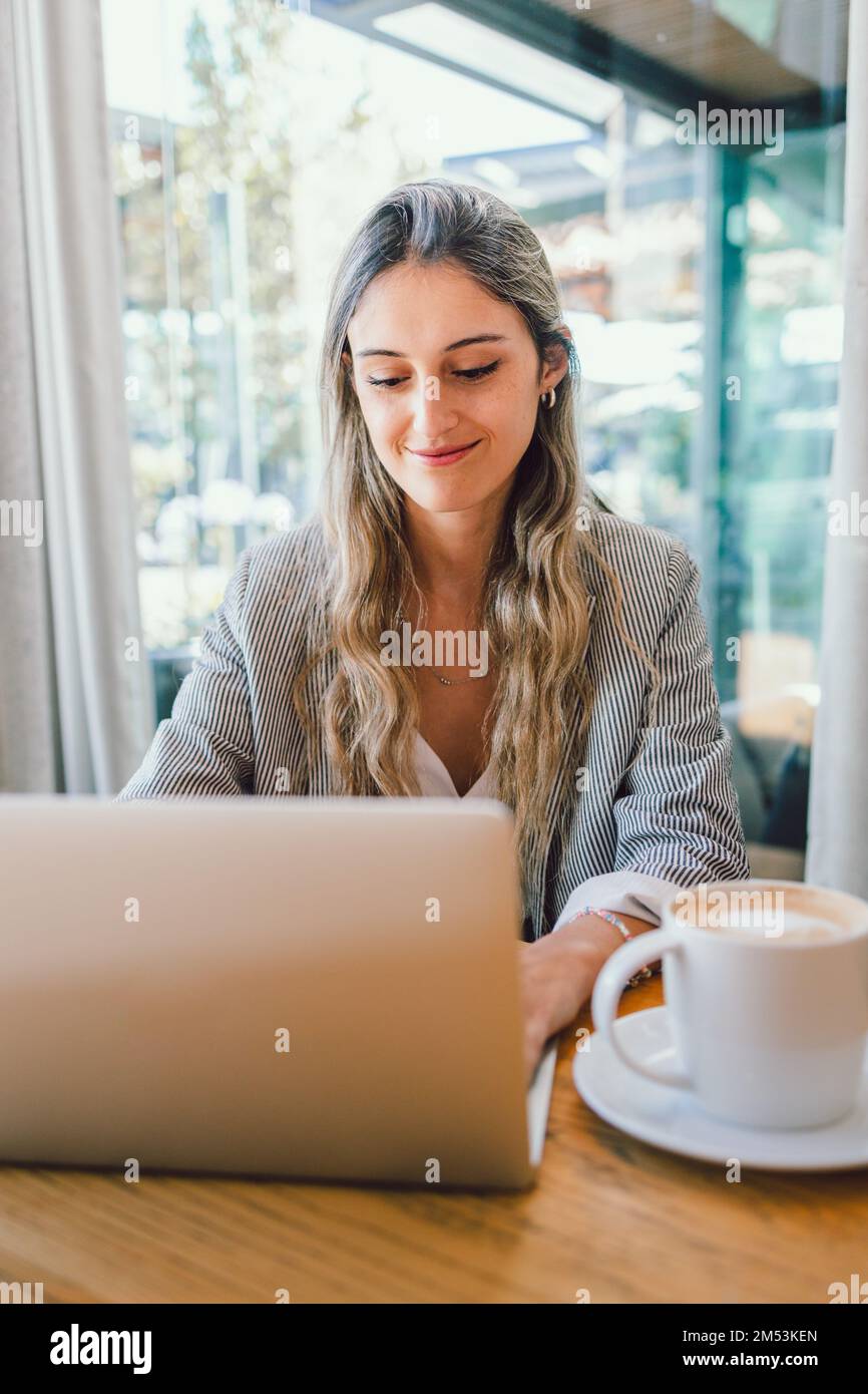 Friedliche Geschäftsfrau, die ein Notebook benutzt, mit einer Tasse Kaffee am modernen Arbeitsplatz. Generation der Millennials Stockfoto
