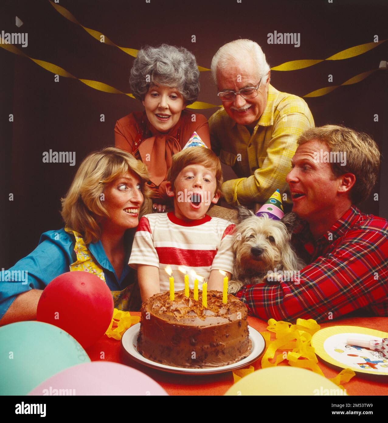 Junge Familie mit Großeltern und Hund schwebte um das Kind herum und blies Kerzen auf seinem Geburtstagskuchen aus Stockfoto