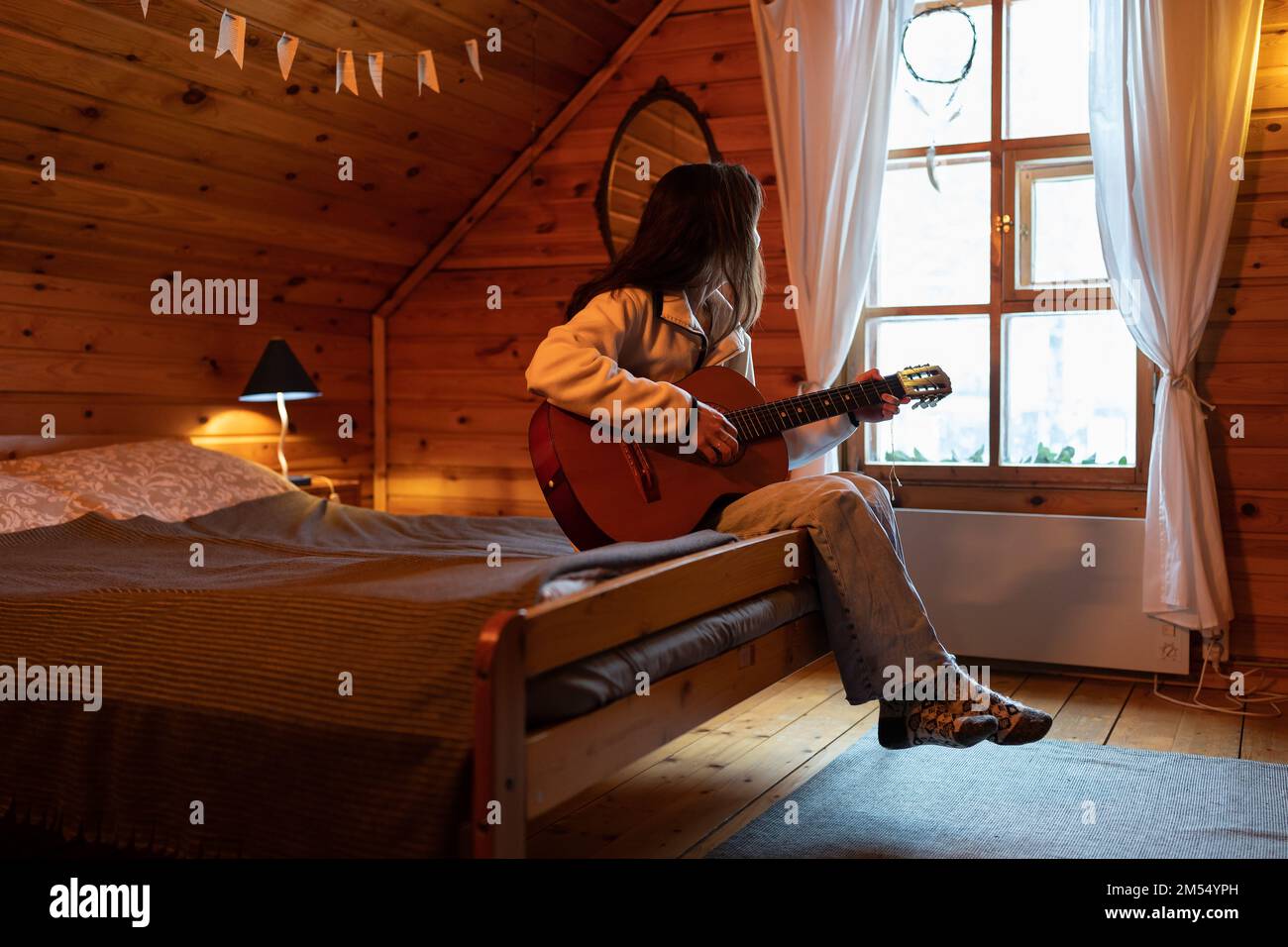 Melancholische einsame Frau sitzt auf dem Bett mit Gitarre in den Händen und schaut aus dem Fenster in einem gemütlichen Holzhaus Stockfoto