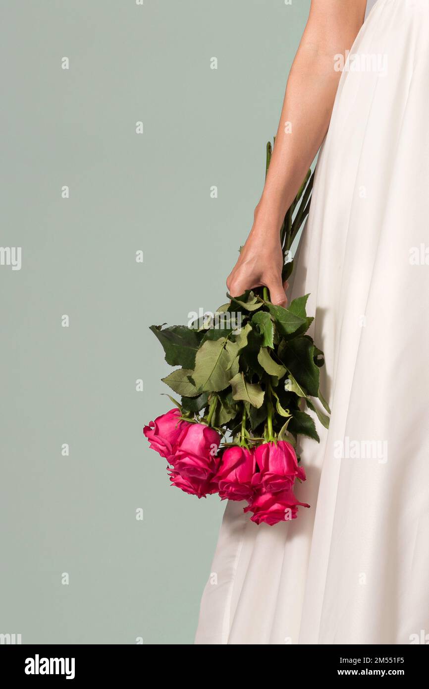Eine anonyme Frau in weißem Kleid mit einem Strauß leuchtend roter Rosen vor grauem Hintergrund am Hochzeitstag Stockfoto