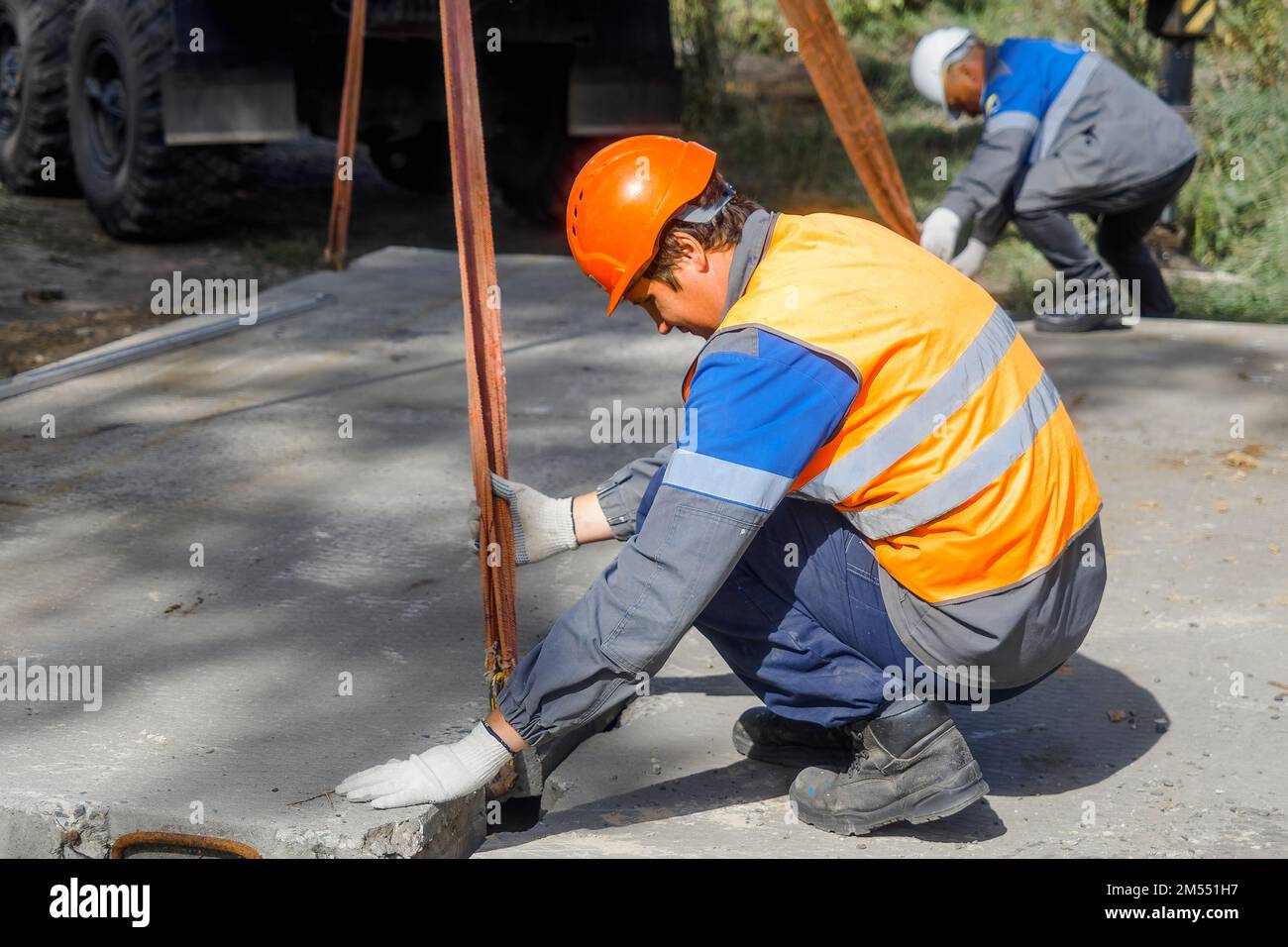 Der Schleuder legt am Sommertag Betonplatten auf die Baustelle. Arbeiter in Schutzweste und Bauhelm beaufsichtigt das Verlegen der Basis auf der Baustelle. Stockfoto