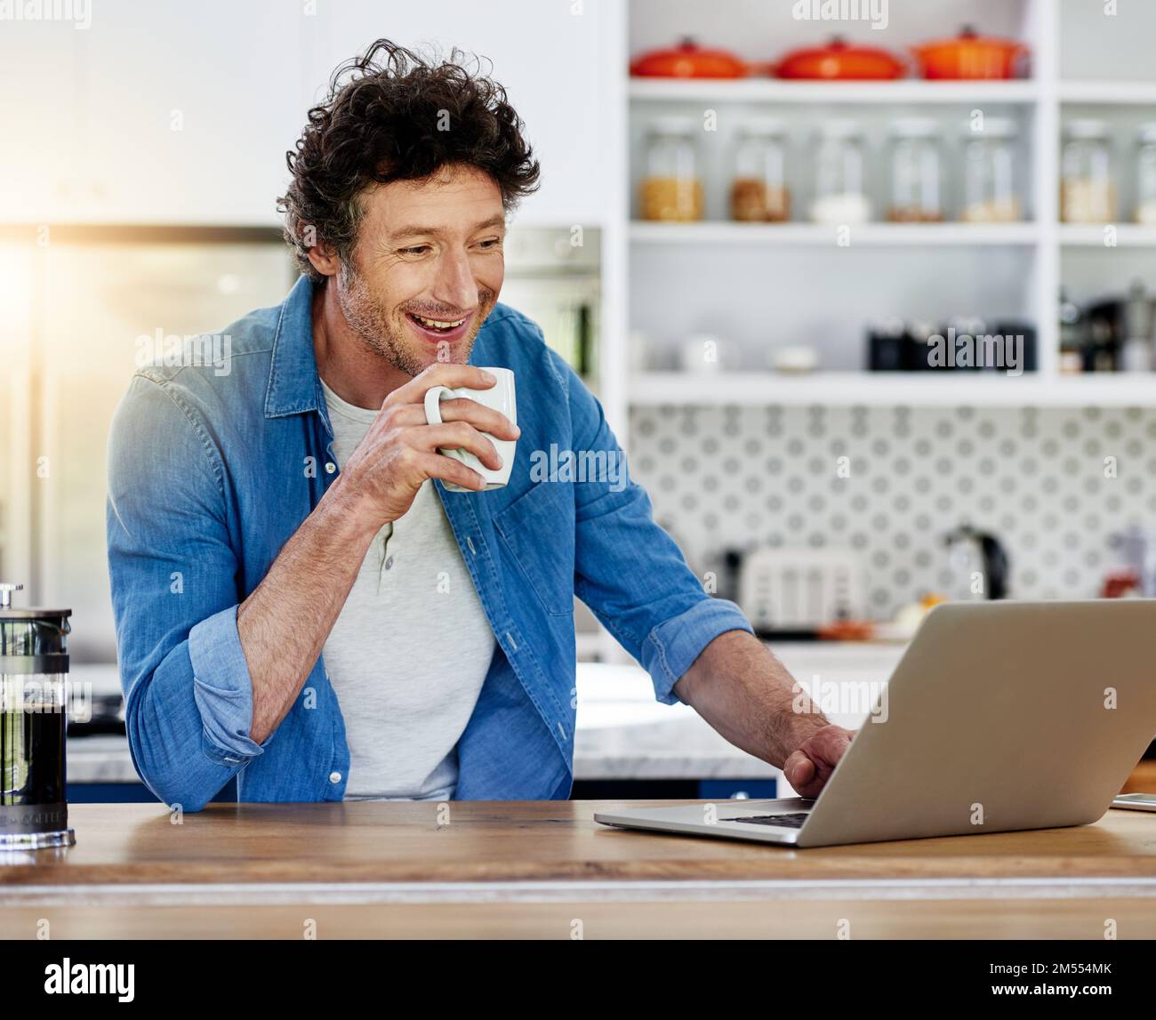 Kaffee und Internet - zwei seiner Lieblingssachen. Ein Junggeselle, der seinen Laptop benutzt, während er eine Tasse Kaffee in der Küche genießt. Stockfoto