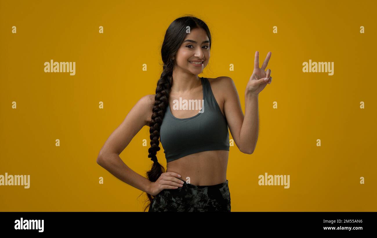 Glückliche indische Sportlerin weibliche Sportlerin im gelben Studiohintergrund zeigt Doppelfinger Geste zwei 2-Nummer Sieg Friedenszeichen Hand Stockfoto