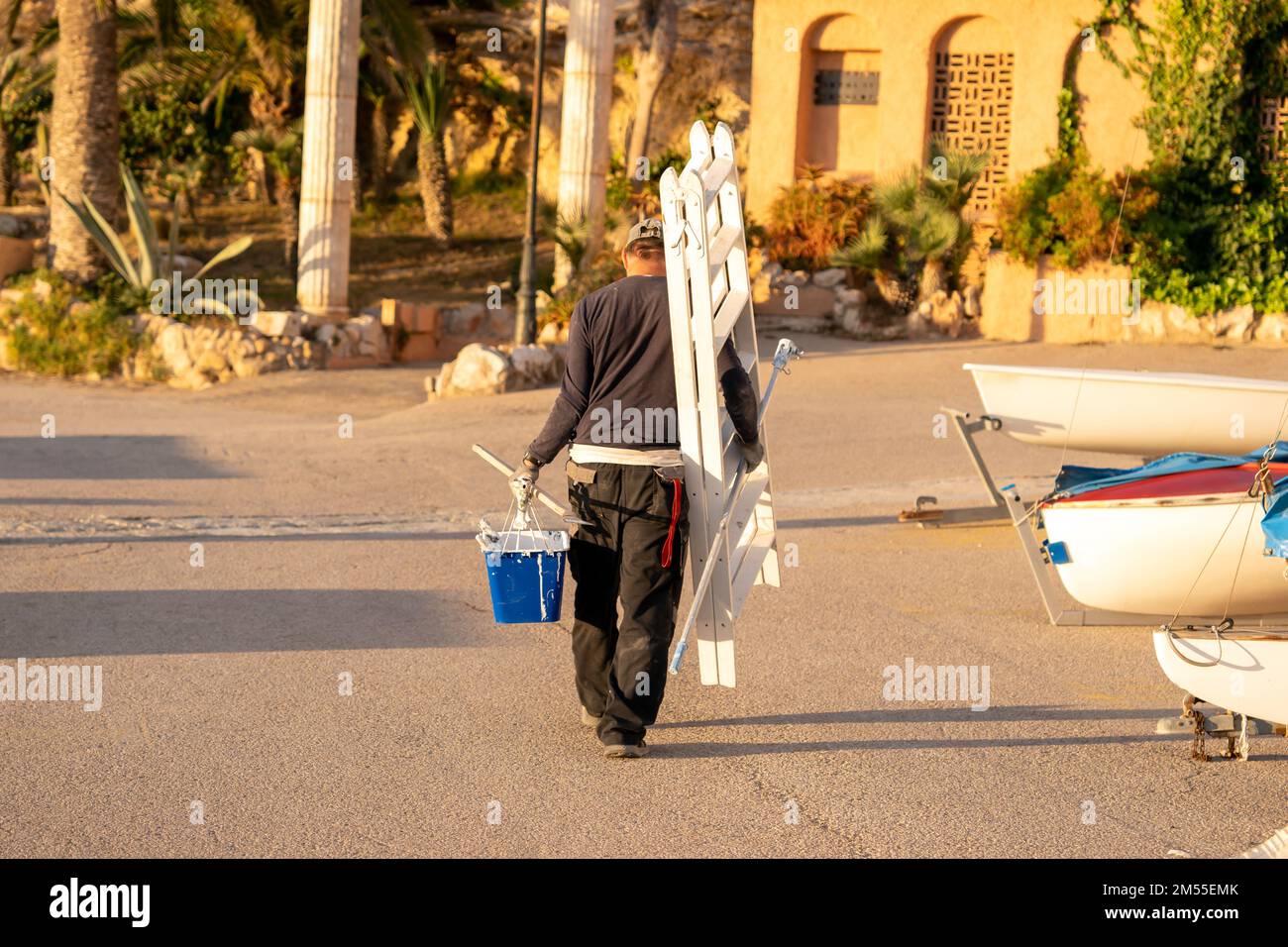 Rückblick auf einen anonymen Angestellten, der eine Leiter und einen Eimer mit weißer Farbe trug, der auf Sand in der Nähe von Booten während der Arbeit am Strand am Morgen lief Stockfoto