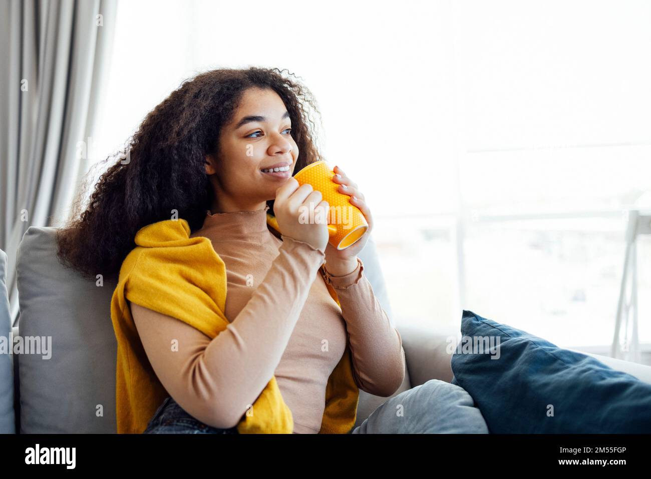 Fröhliches, lächelndes afroamerikanisches Mädchen, das sich auf der Couch mit einer Tasse Kaffee entspannt, sich zu Hause ausruht, den freien Tag genießt, warme Kleidung trägt Stockfoto