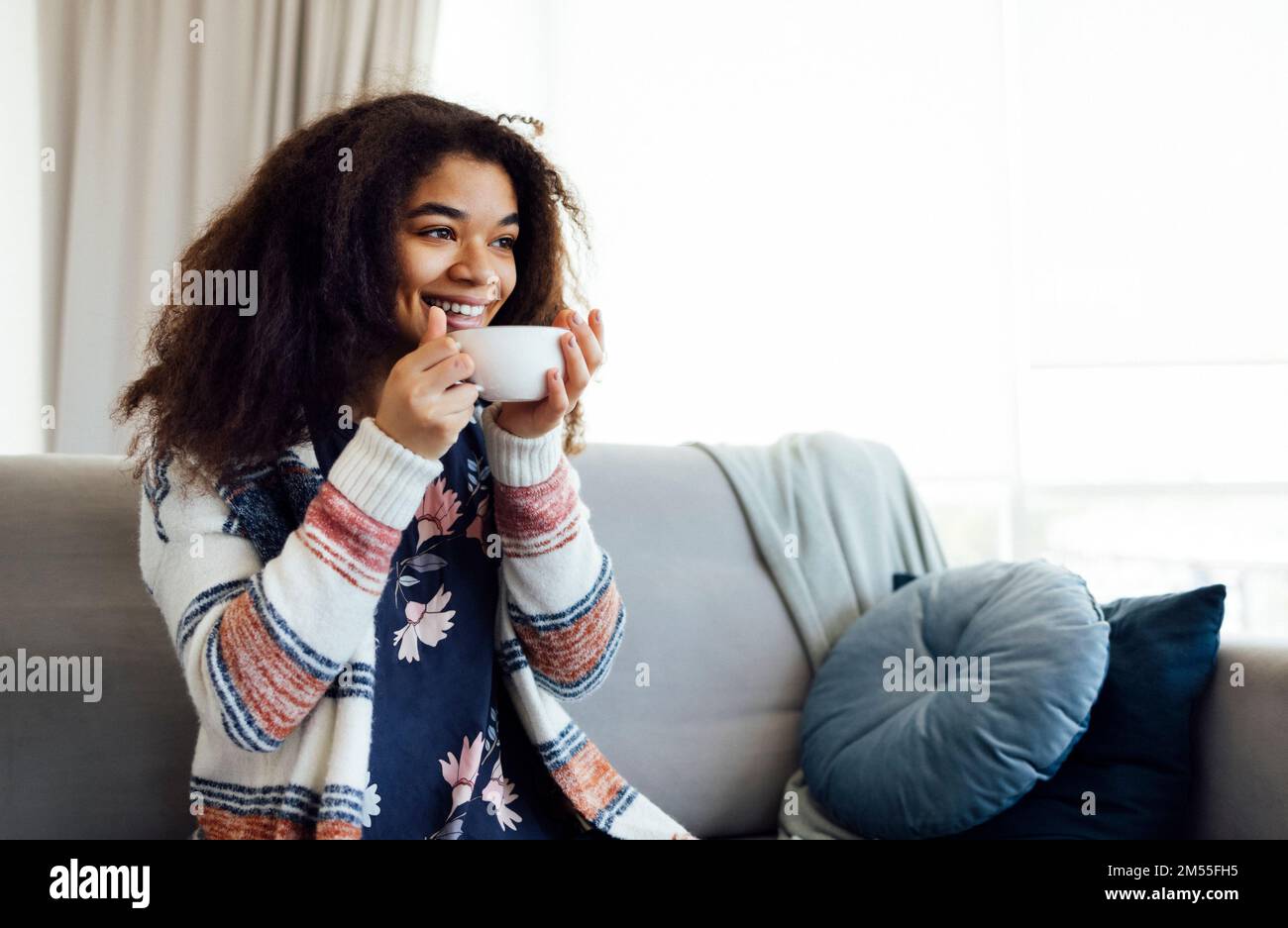 Fröhliches, lächelndes afroamerikanisches Mädchen, das sich auf der Couch mit einer Tasse Kaffee entspannt, sich zu Hause ausruht, den freien Tag genießt, warme Kleidung trägt Stockfoto