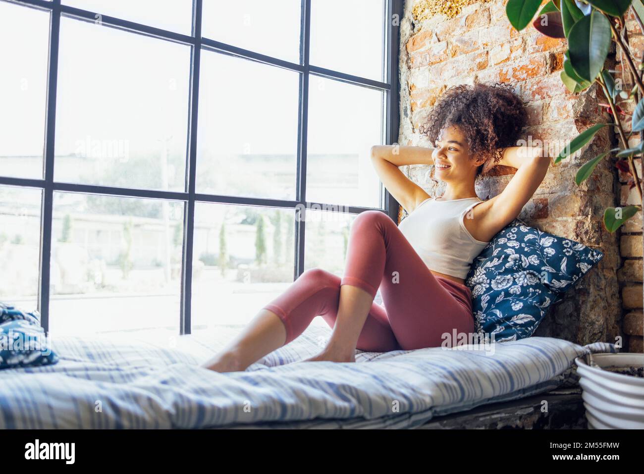 Ein entspanntes, afroamerikanisches Mädchen in Hausbekleidung, das auf einer Fensterbank sitzt, eine glückliche Afro-Frau zu Hause, die sanft zum Fenster lächelt Stockfoto Ein entspanntes, afroamerikanisches Mädchen in Hausbekleidung, das auf einer Fensterbank sitzt, eine glückliche Afro-Frau zu Hause, die sanft zum Fenster lächelt Stockfoto