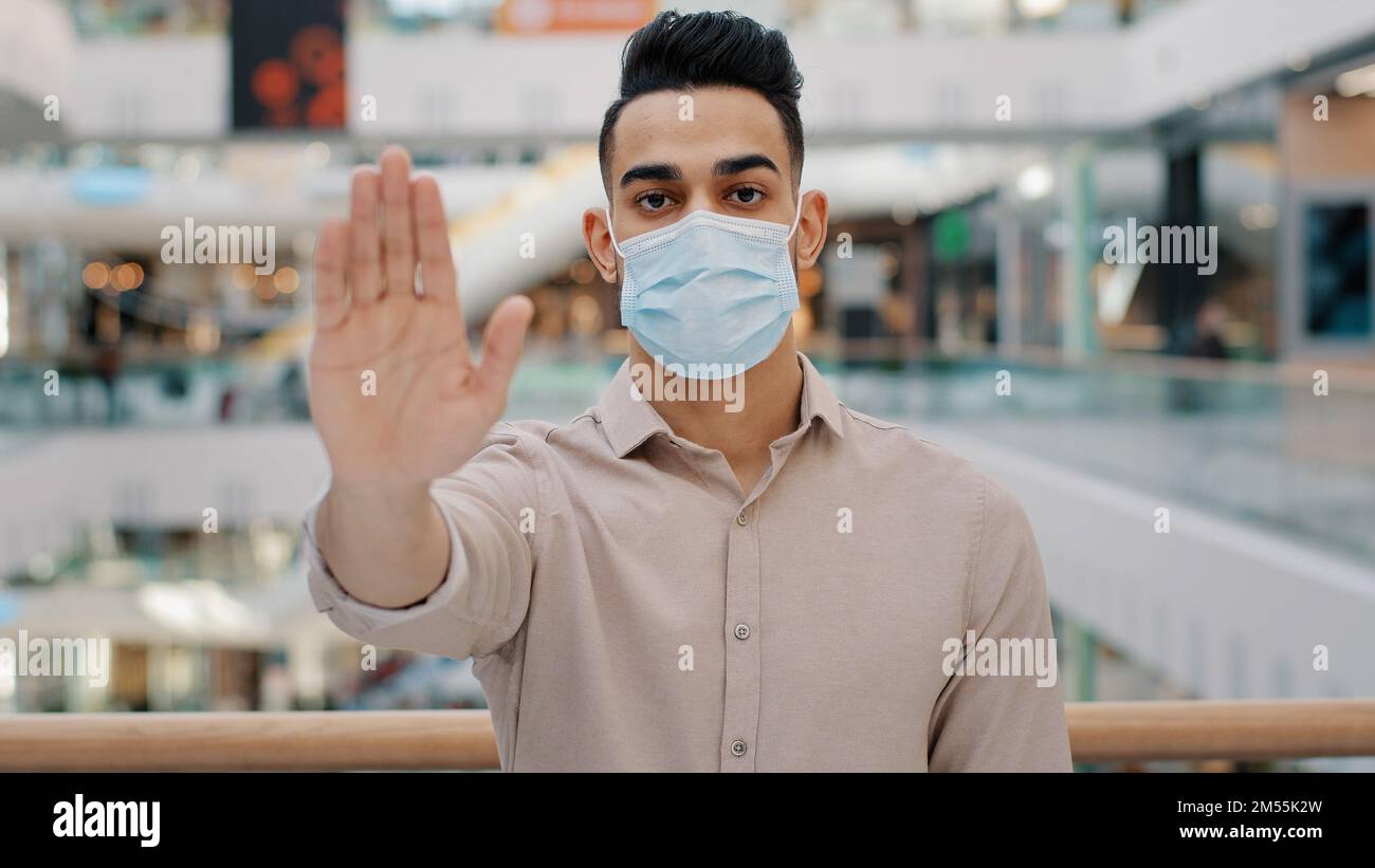 Portrait des jungen hispanischen Mannes, der eine medizinische Maske trägt, Handfläche nach vorne legen ernsthaften ethnischen Kerl, der auf Kamera Männchen schaut, zeigt Stop-Zeichen-Handfläche mit Hand Stockfoto
