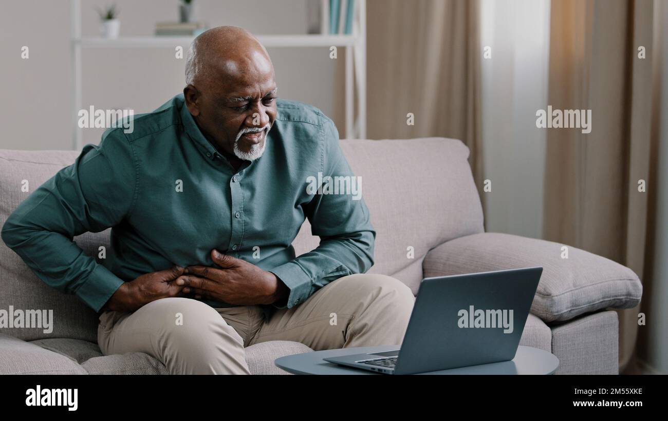 Älterer afroamerikanischer Freiberufler reifer Rentner, der im Heimbüro auf der Couch sitzt und einen Laptop benutzt, fühlt akute Magenschmerzen Unwohlsein Verdauungsstörungen Stockfoto
