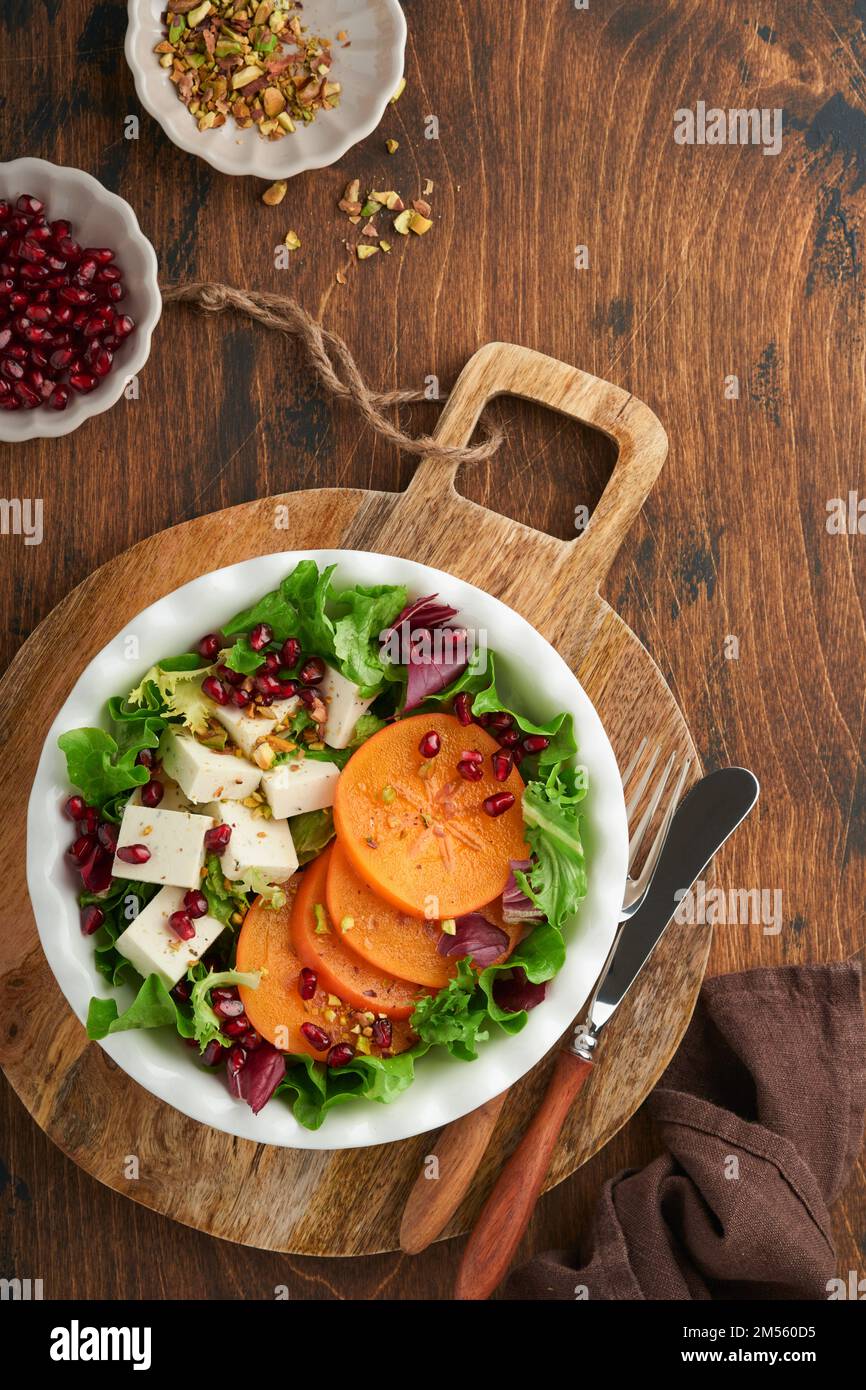 Salat. Frischer grüner Salat mit Fetakäse, Persimone, Granatapfel und Pistazien auf weißem Teller. Eine Idee für einen gesunden, köstlichen Winter-Weihnachtssalat. - Nein Stockfoto