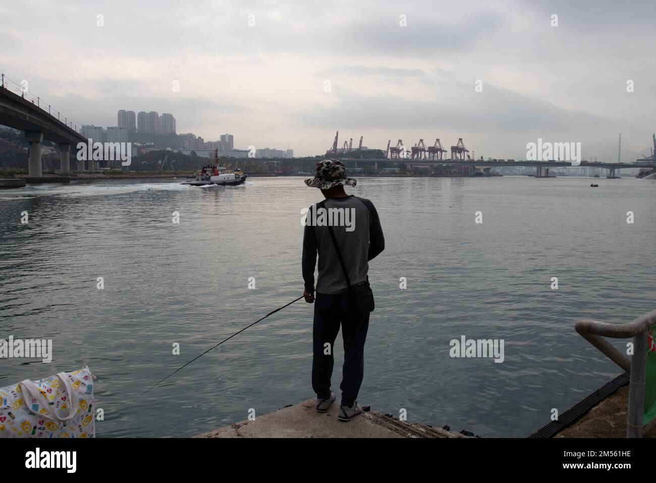 Ein Mann mit einem Floppy-Eimer-Hut beim Angeln mit einer Angelrute im Meer, der auf dem Dock in der Nähe der Brücke steht Stockfoto