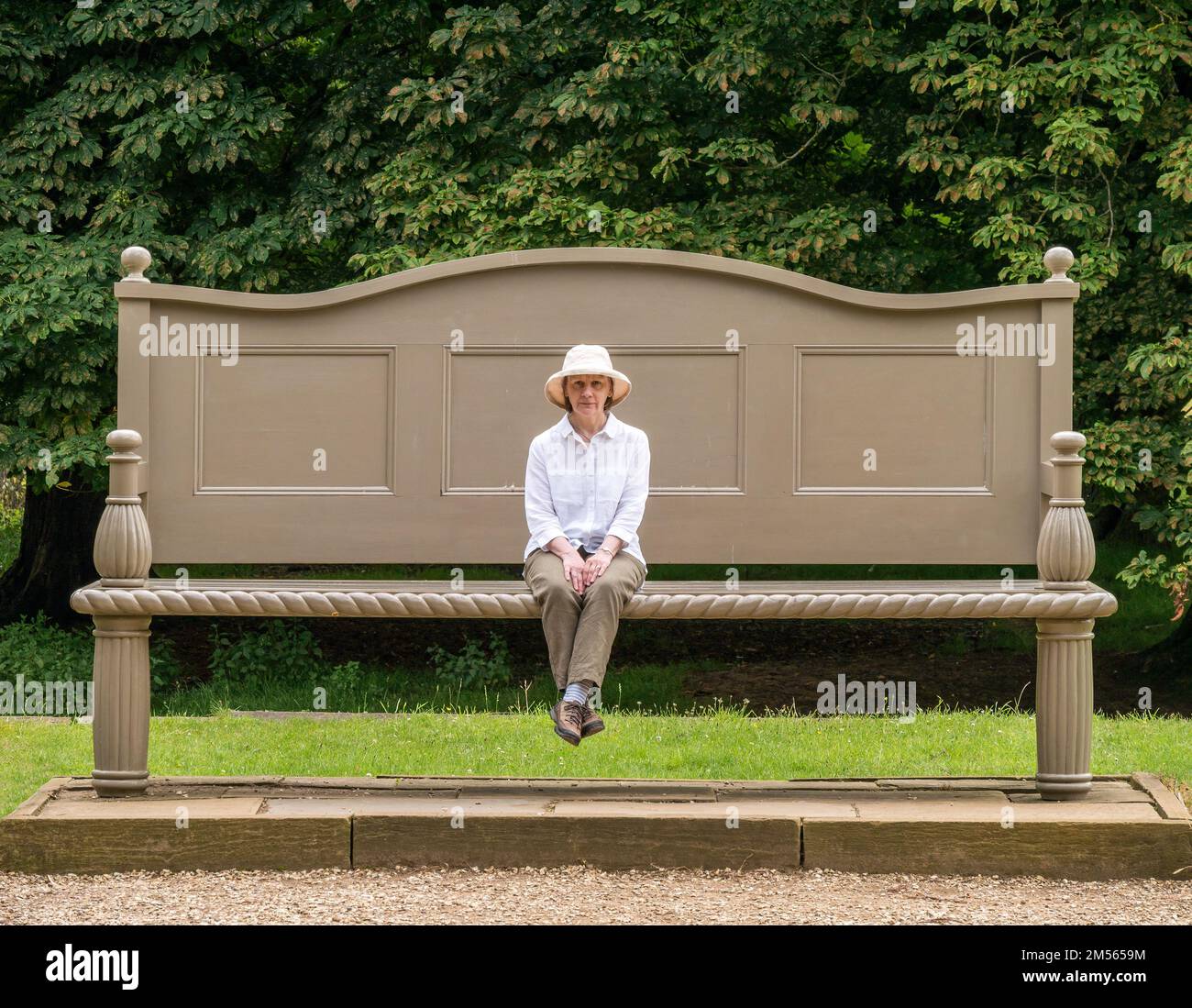 Illusion, kleine Frau, die auf einer übergroßen Gartenbank in Belton House Garden Grounds, Grantham, Lincolnshire, Großbritannien sitzt Stockfoto