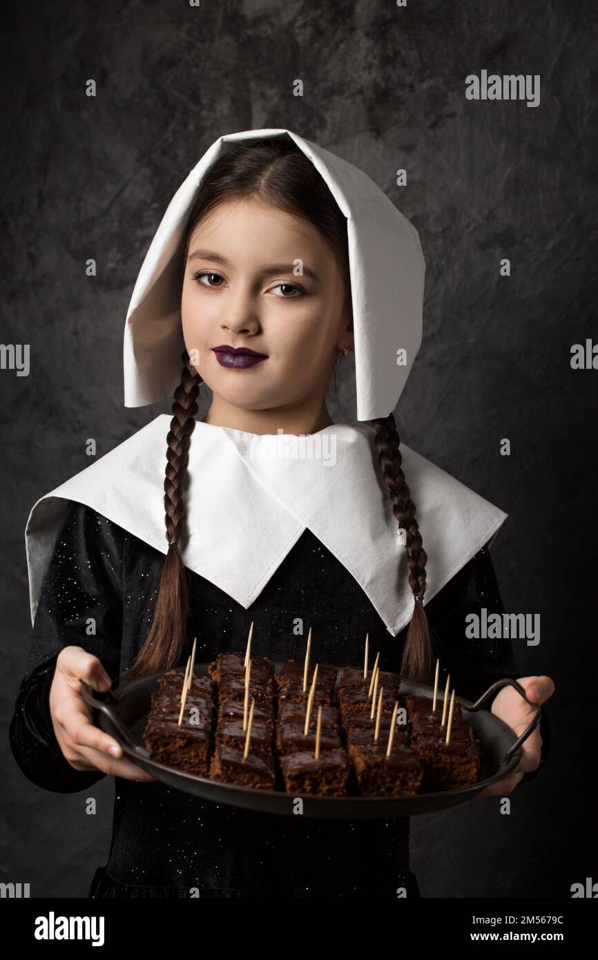 Mittwoch Addams. Gotisches Mädchen mit Zöpfen in einer Mütze hält ein Tablett mit Schokoladenkuchen auf dunklem Hintergrund Stockfoto