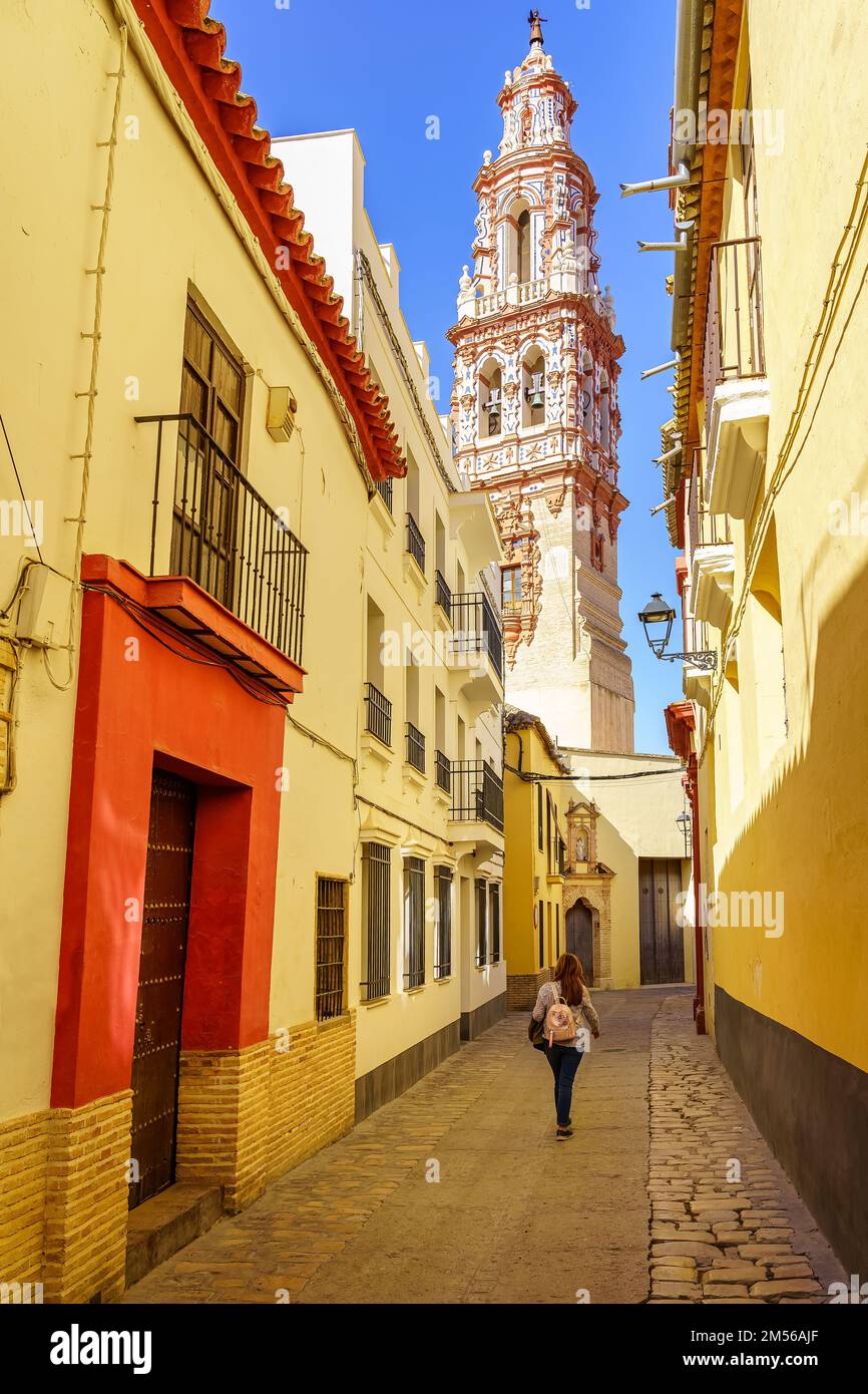 Frau, die durch die engen Gassen der Stadt Ecija spaziert, mit dem beeindruckenden Kirchturm im Hintergrund. Stockfoto