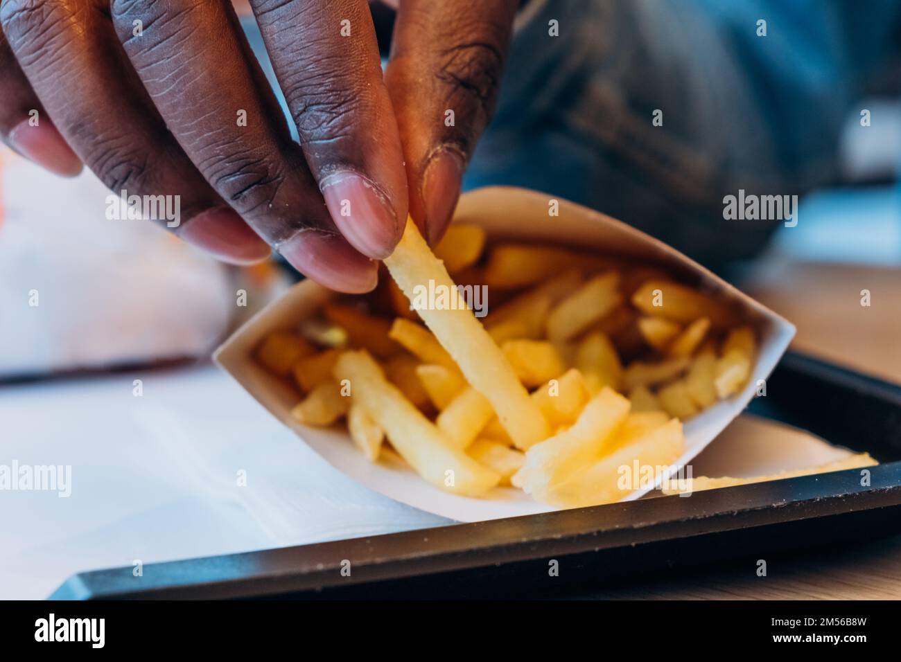 Ein junger afroamerikanischer Mann nimmt leckere frische pommes aus dem Papierbehälter an den Tisch im modernen Café, ganz aus der Nähe Stockfoto