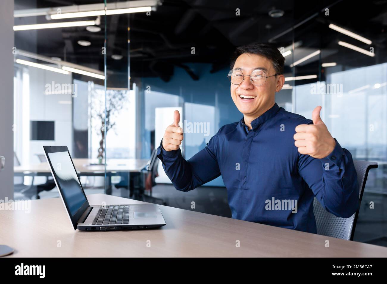 Porträt eines jungen, gutaussehenden Mannes, asiatischer Designer, Ingenieur, Freiberufler. Er sitzt mit einem Laptop im Büro am Tisch, schaut in die Kamera, lächelt, zeigt seine Hände mit seinen Fingern super. Stockfoto