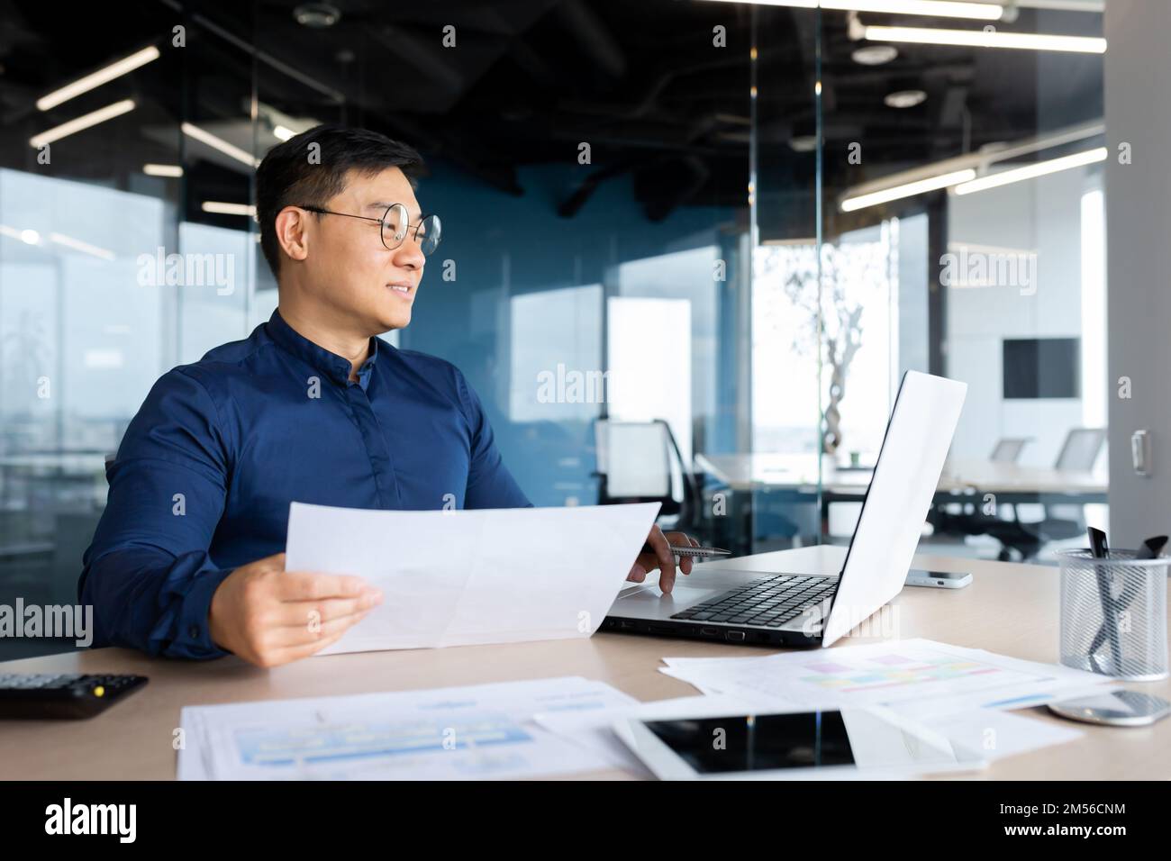 Ein junger asiatischer Journalist, Blogger, Freiberufler sitzt im Büro am Tisch. Funktioniert auf einem Laptop und mit Papieren. Schreibt einen Artikel, ein Buch, ein Interview. Stockfoto