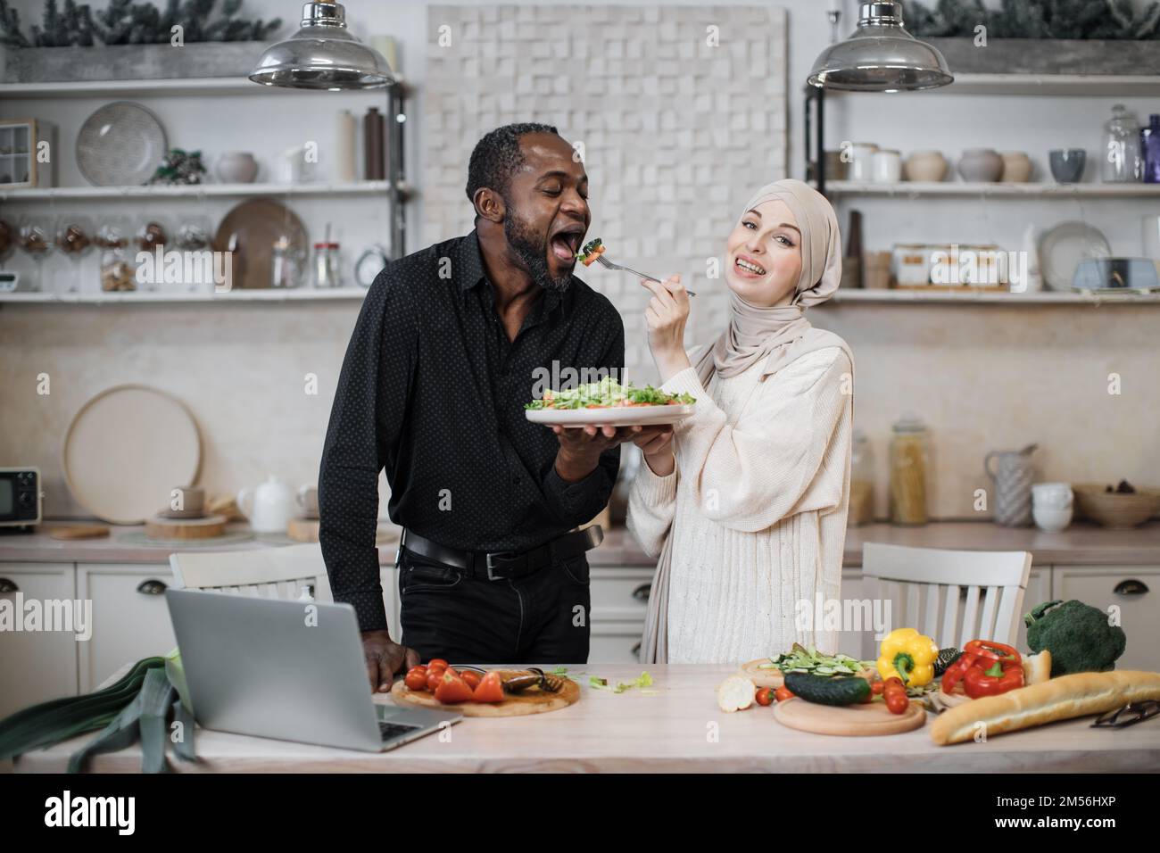 Fröhliches, multinationales Ehepaar, das ein Notebook benutzte, während es gesundes Essen in der Küche kochte, junge muslimische Frau im Hijab fütterte ihren Mann mit einer Gabel mit Salat aus frischem Gemüse. Stockfoto